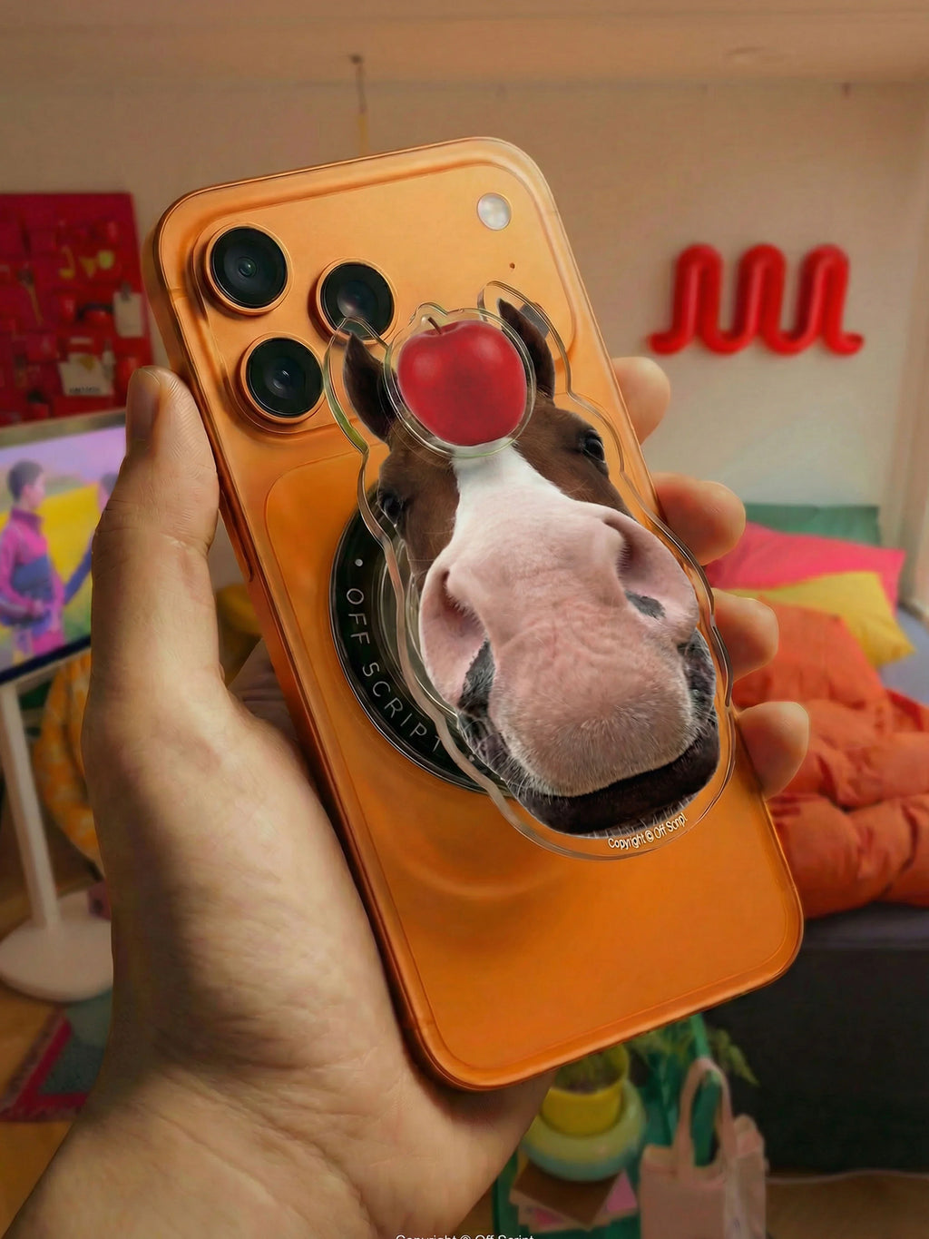 Close-up of OFF SCRIPT Apple-Horse MagSafe pop-up grip on orange iPhone, showing horse face holding a red apple